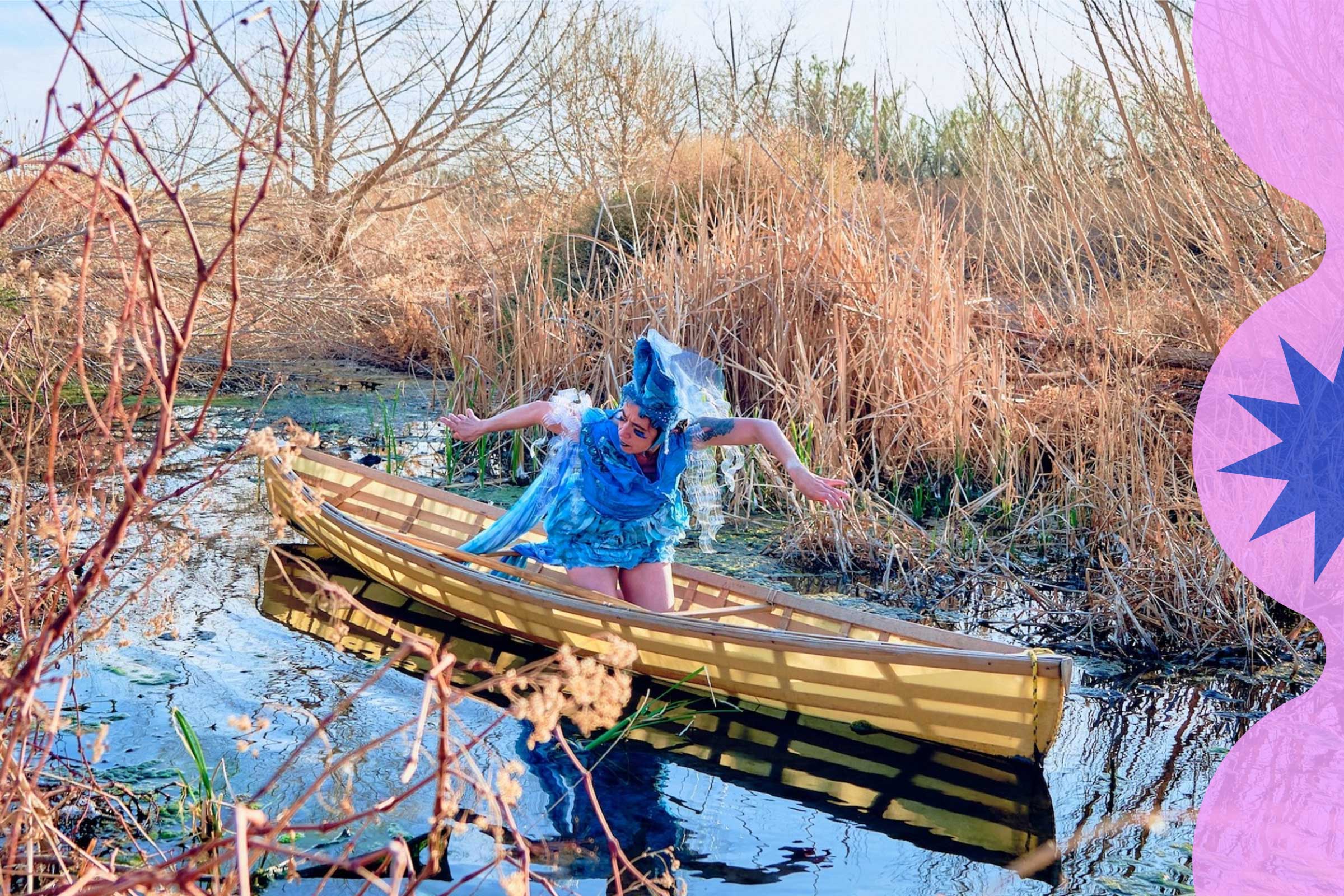 A dancer in blue poses in a canoe in a body of water surrounded by tall rushes and reeds.