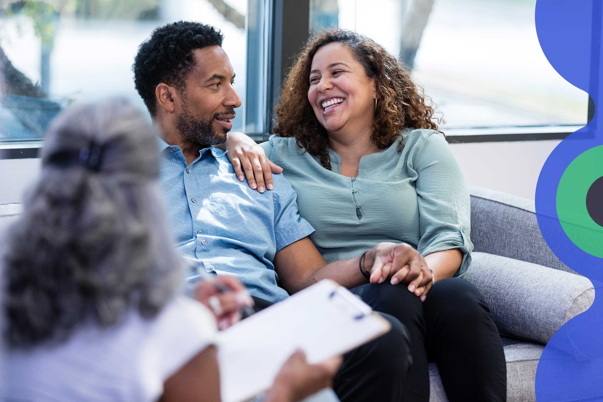A couple sit, laughing, on a couch together in a windowed room across from a woman with a clipboard whose back is to the camera.